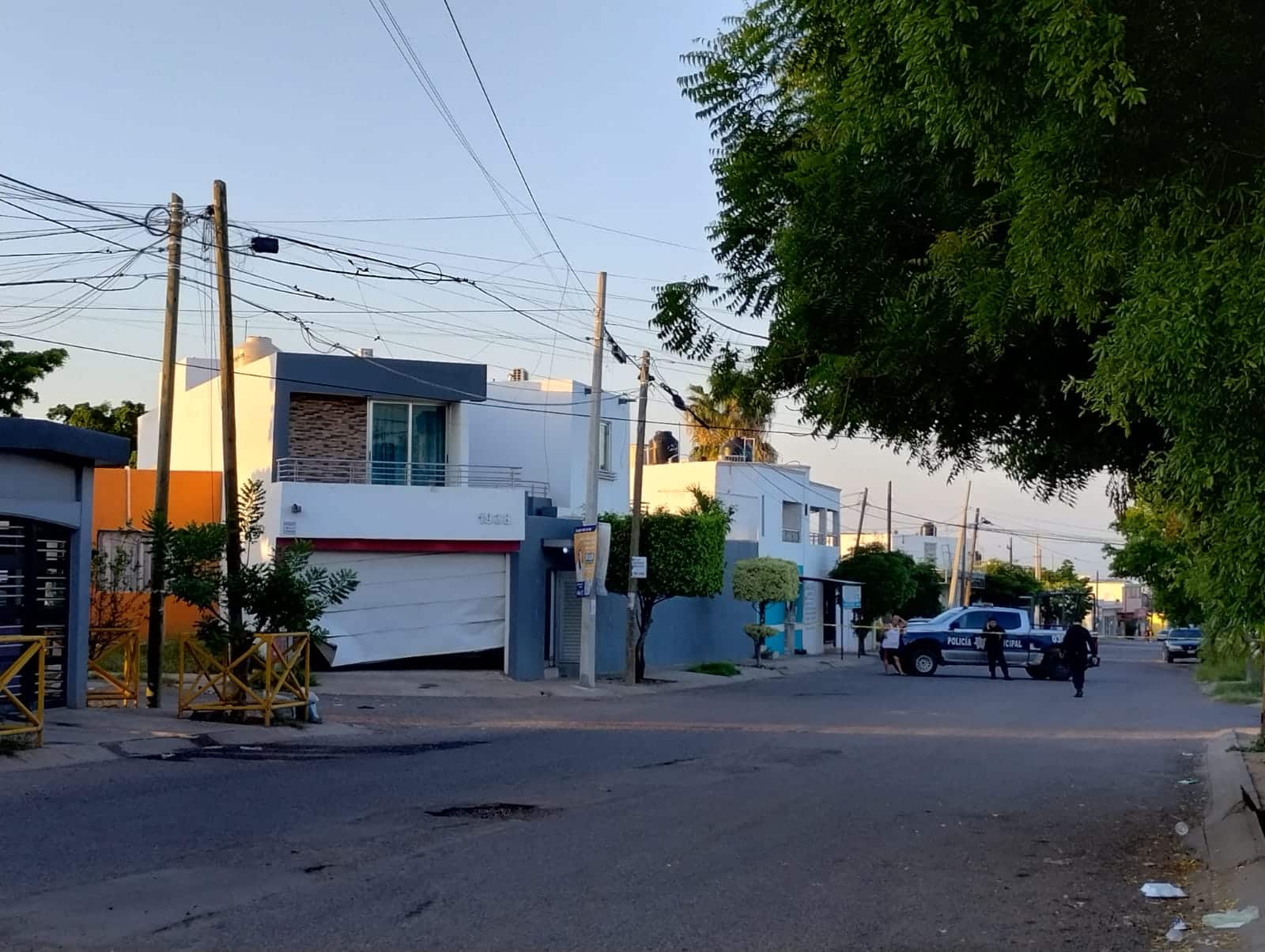 Personas, árboles, calle pavimentada, casas y una patrulla de la policía municipal