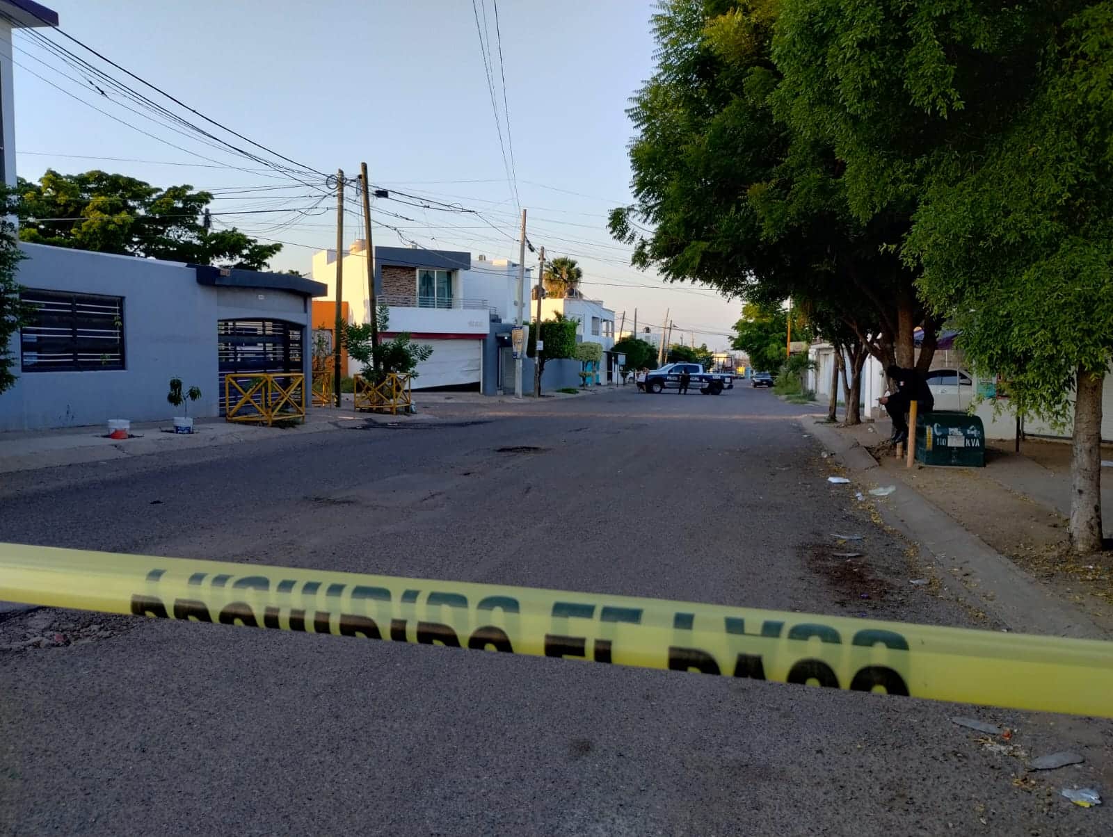 Cinta amarilla delimitando el área donde le dispararon a una persona, árboles, calle pavimentada, casas, personas y una patrulla de la policía municipal