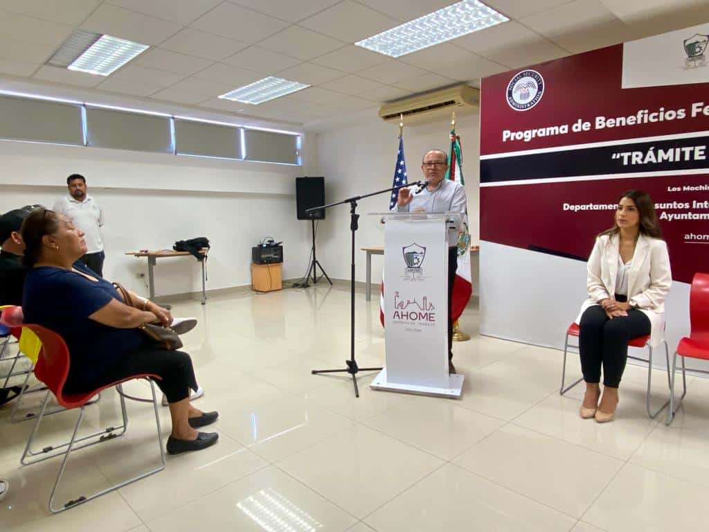 Personas sentadas y una de pie en un podium, un micrófono y atrás banderas