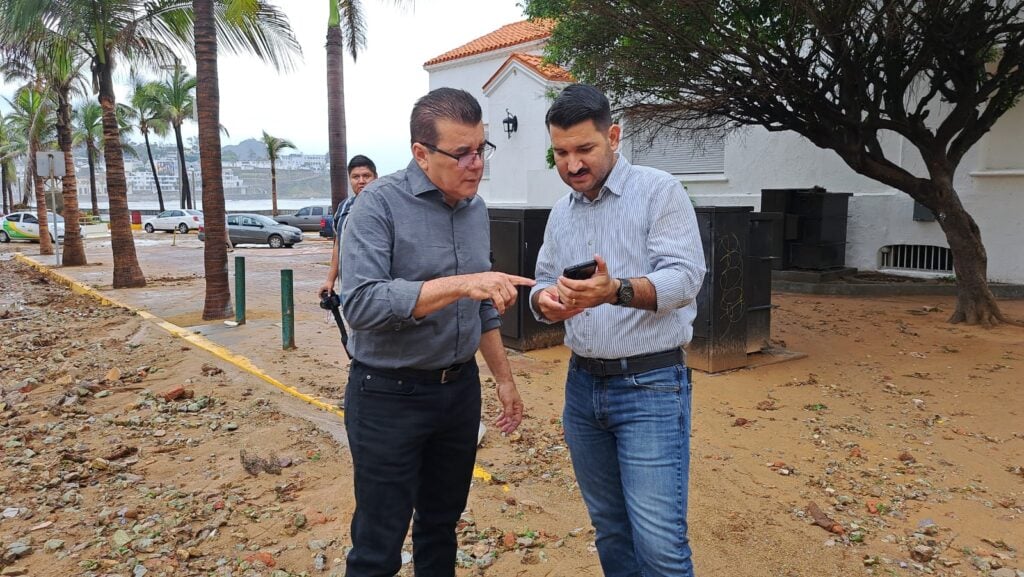 2 personas mirando un celular, una calle con piedras obstruyendo el paso, palmeras, carros, casas y al fondo el mar