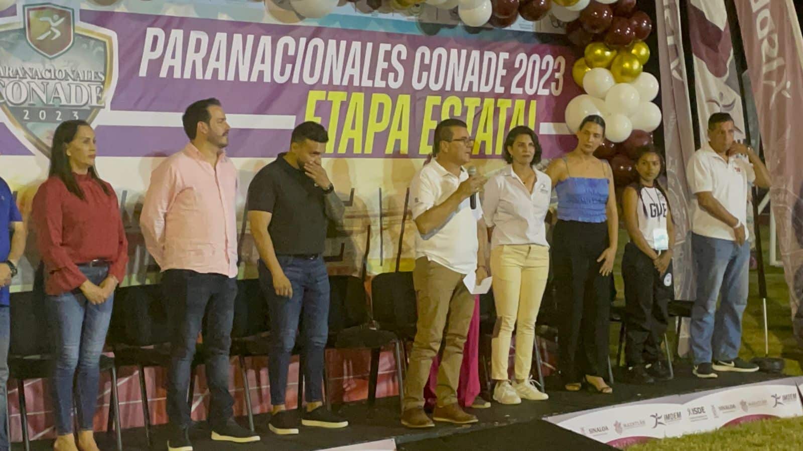 Personas de pie, sillas, globos y una lona al fondo