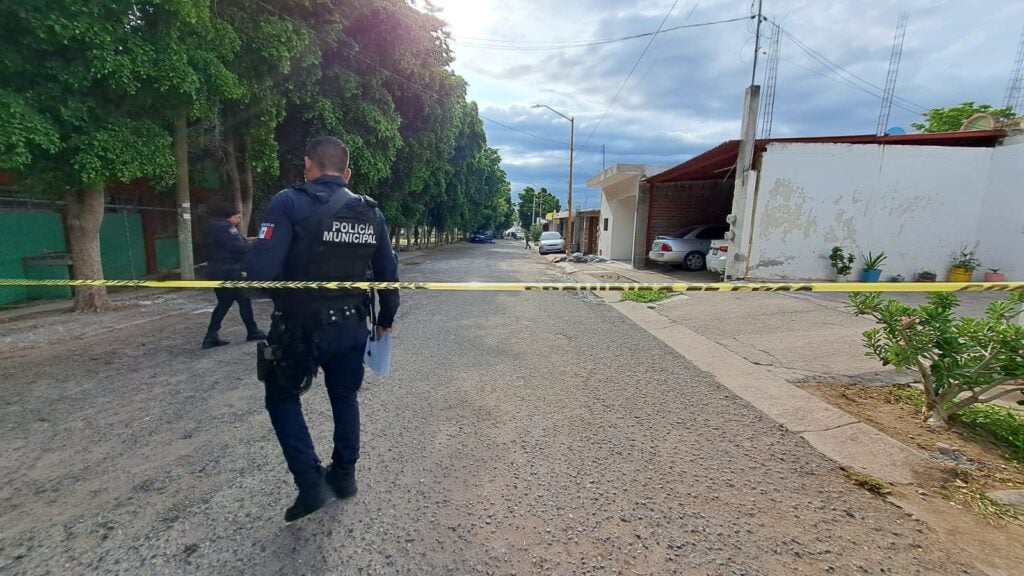 2 personas con vestimenta de la Policía Municipal, carros, cinta amarilla, casas y árboles