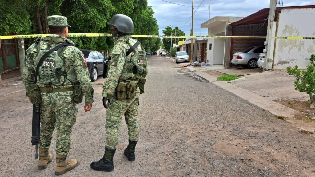 2 personas con vestimenta del Ejército Mexicano, carros, cinta amarilla y árboles