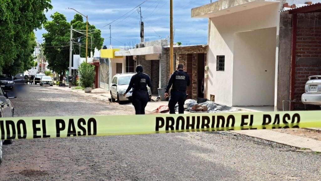 2 personas con vestimenta de la Policía Municipal, carros, cinta amarilla, casas y árboles