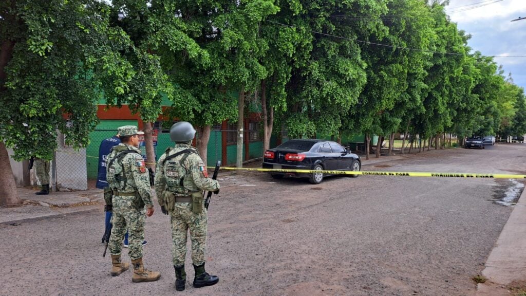 2 personas con vestimenta del Ejército Mexicano, carros, cinta amarilla y árboles