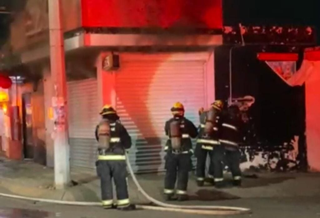 4 personas con traje de bomberos tratando de abrir las cortinas de un expendio al que le sale humo