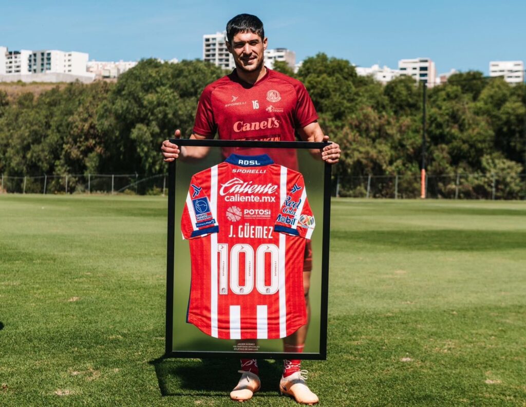Una persona con uniforme de futbol, con un cuadro que enmarca una camisa con el número 100 en una cancha