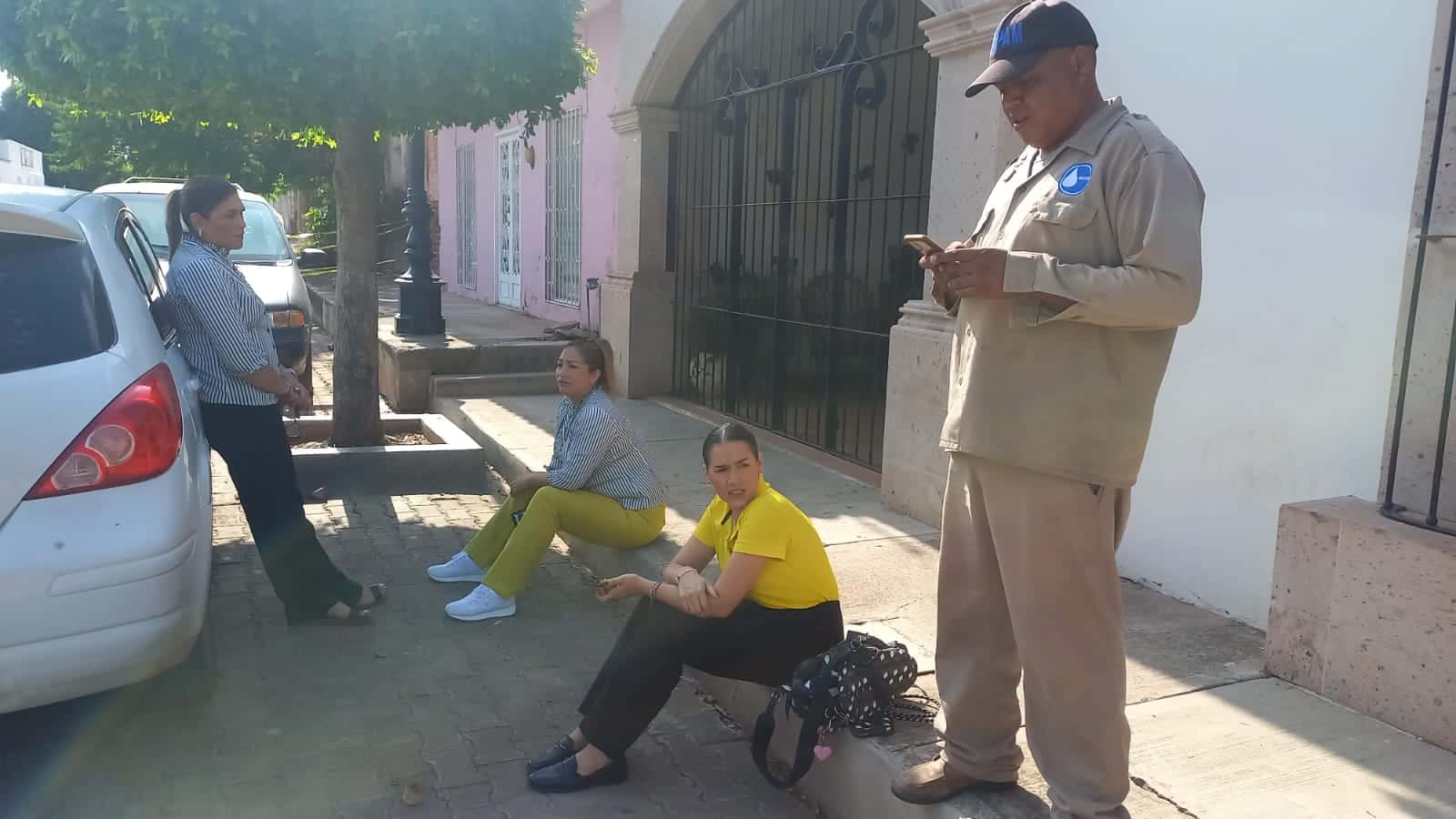 3 personas sentadas y una de pie, árboles, carros estacionados y casas