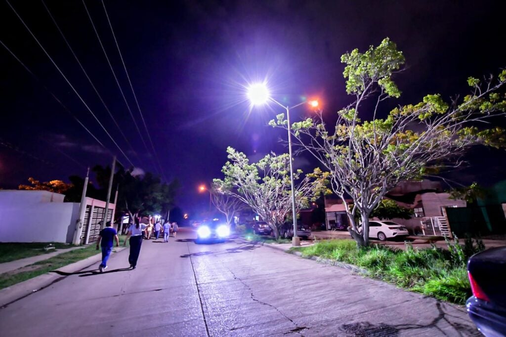 Personas caminando en una calle, carros y una lámpara alumbrando