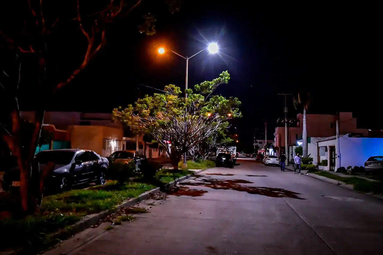 Personas caminando en una calle, carros y una lámpara alumbrando