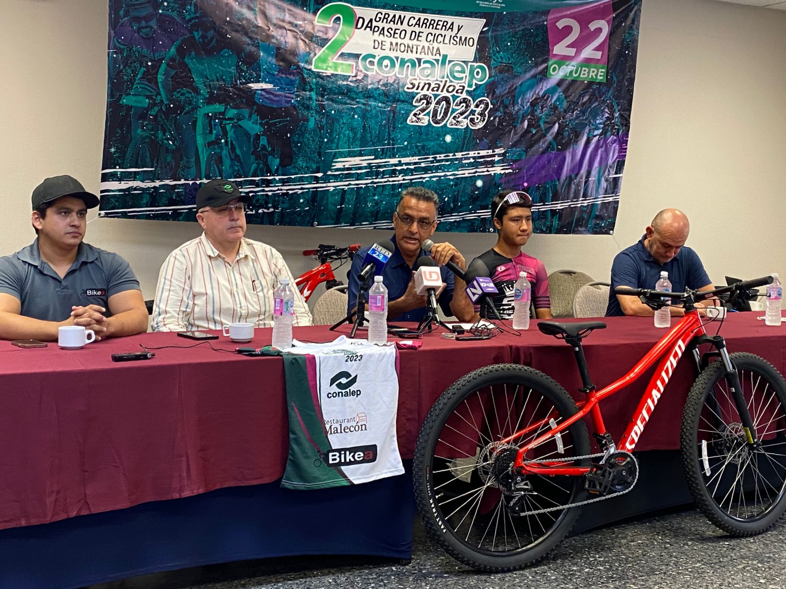 5 personas sentadas, una mesa con mantel guinda, una playera y una bicicleta