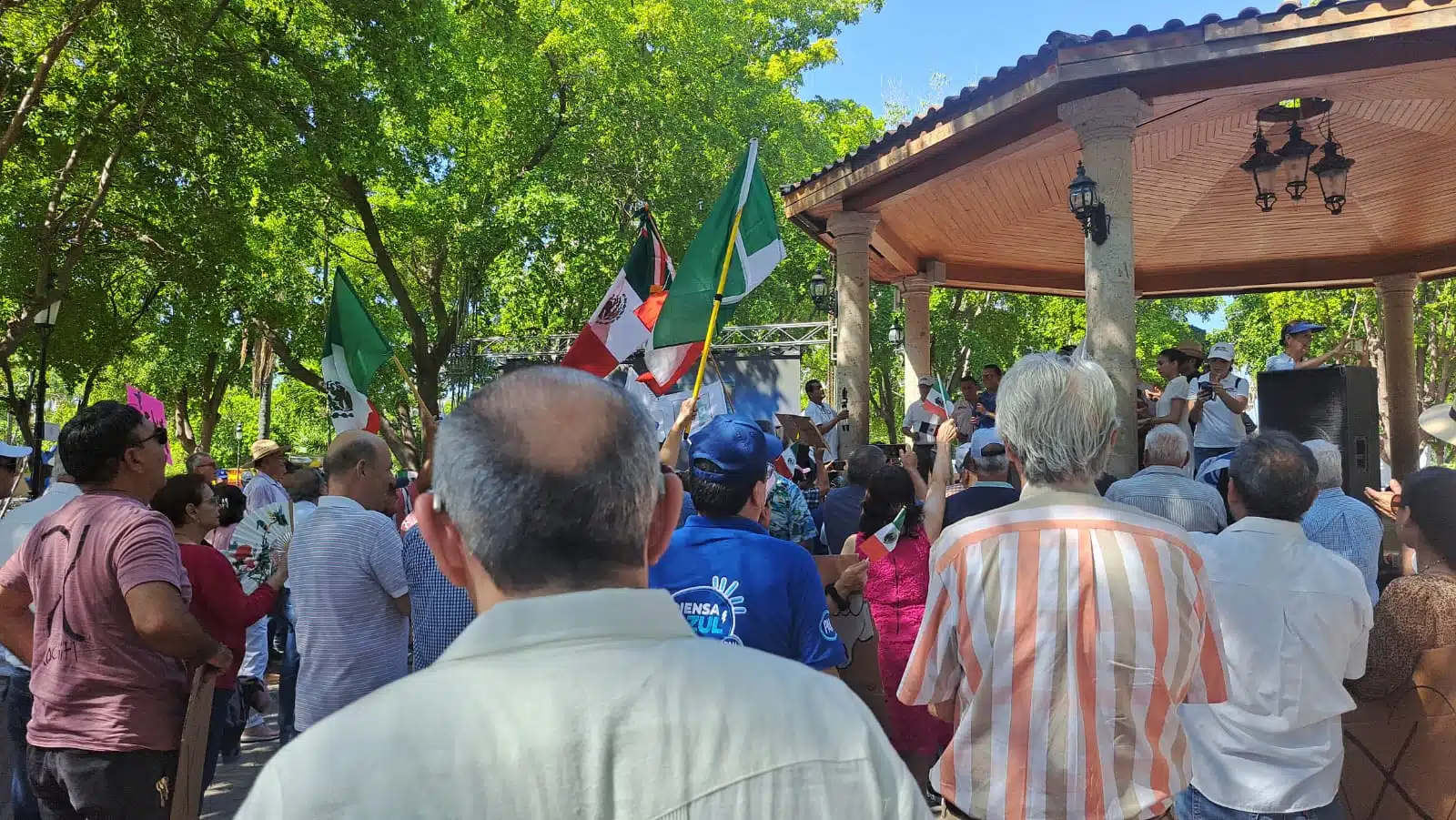 Personas con banderas de México, del PRI y PAN, árboles y parte de un kiosko