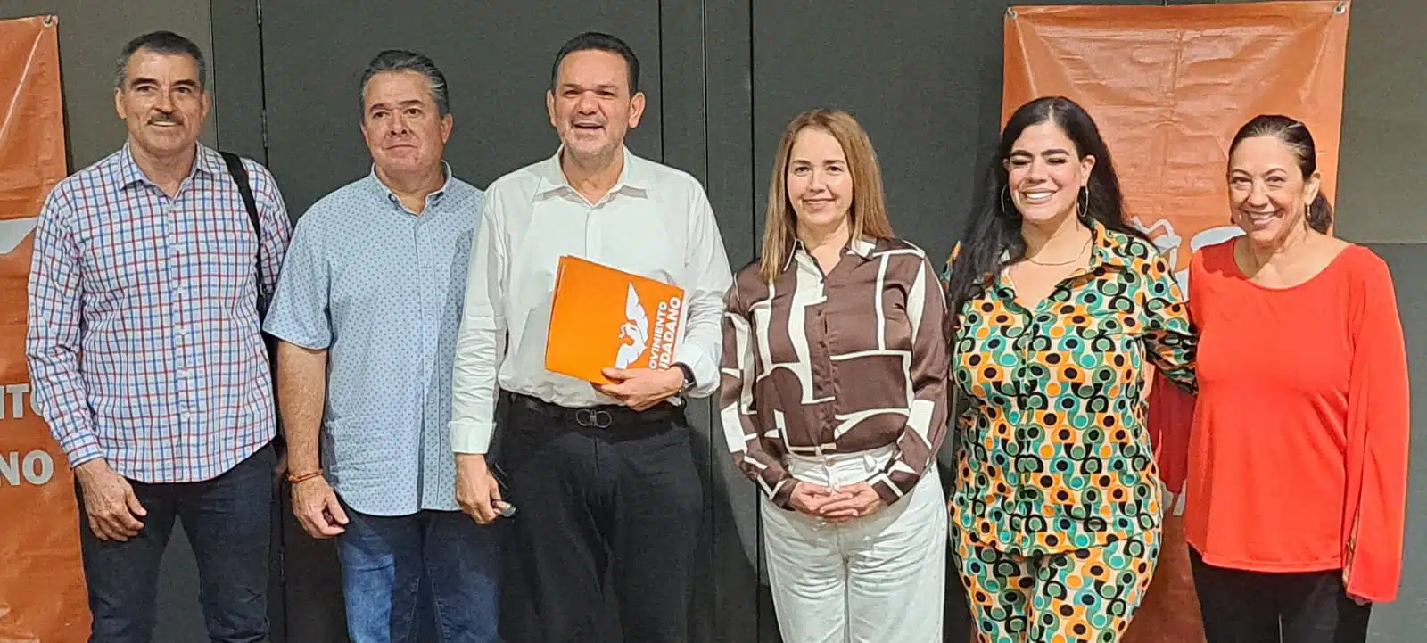 6 personas de pie posando para una foto y una de ellas con una carpeta color naranja