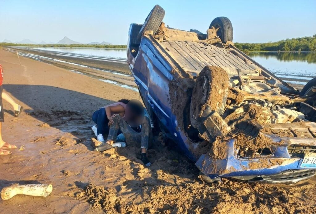 Un carro volcado por una calle de terracería y personas al lado de él