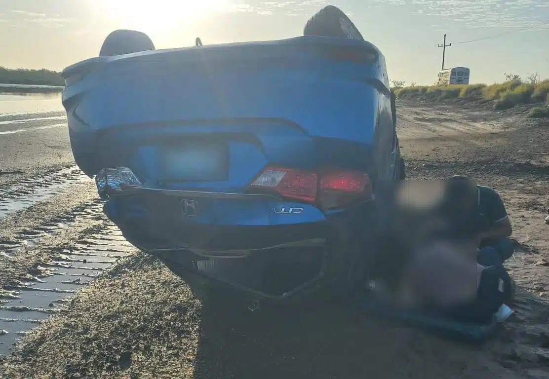 Un carro volcado por una calle de terracería y personas al lado de él