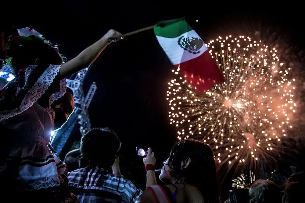 Personas celebrando el Grito de Independencia con fuegos artificiales y una bandera de México