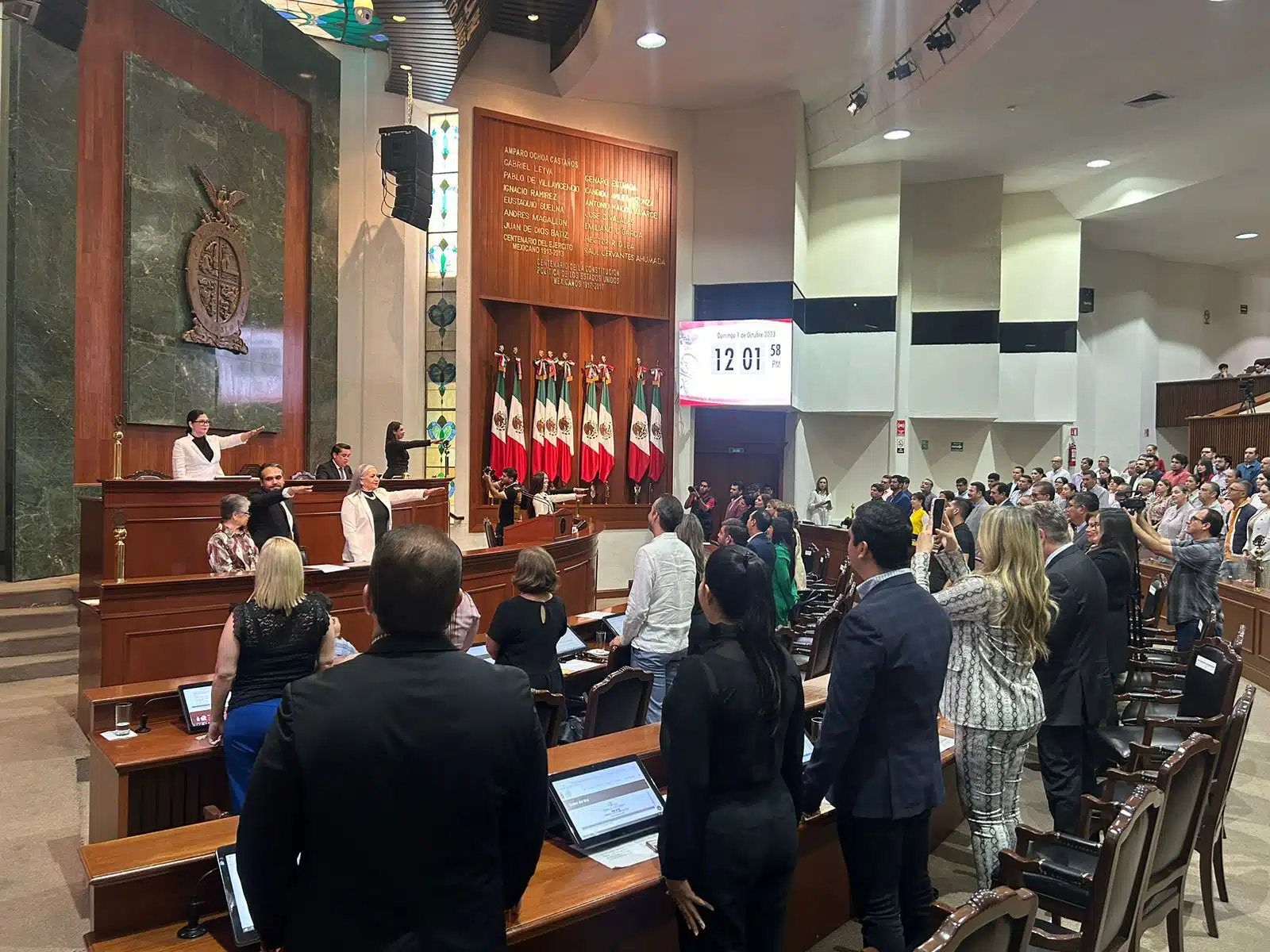 Personas de pie, algunas con la mano levantada en forma de tomando protesta y banderas de México