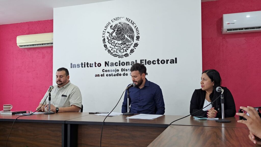 3 personas sentadas, micrófonos de los medios de comunicación y al fondo el sello de la bandera de México