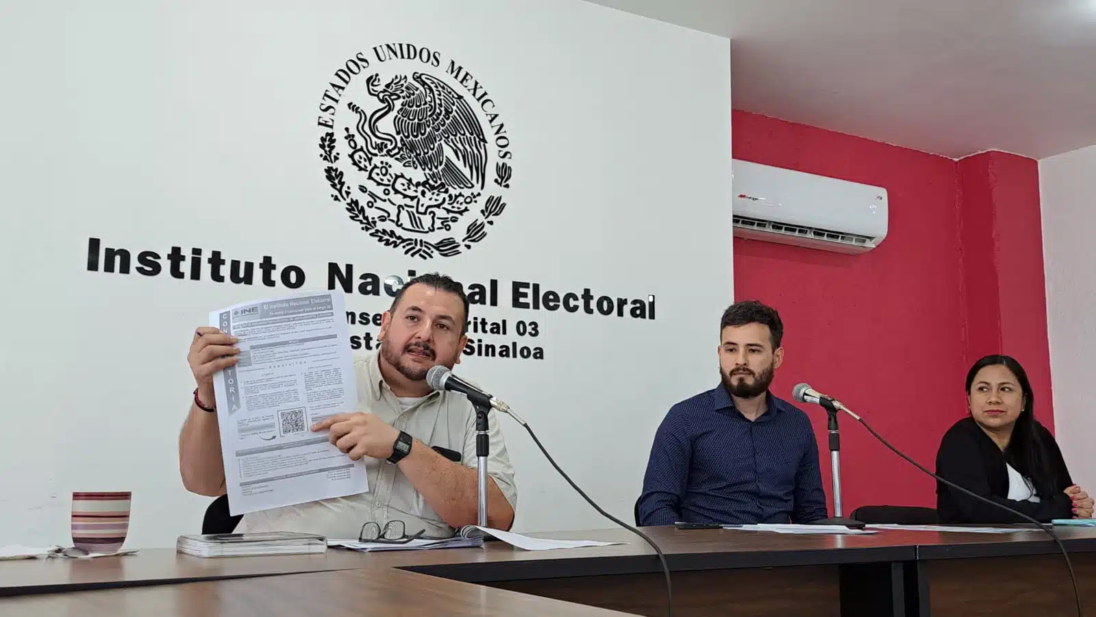 3 personas sentadas, una de ellas hablando, micrófonos de los medios de comunicación y al fondo el sello de la bandera de México