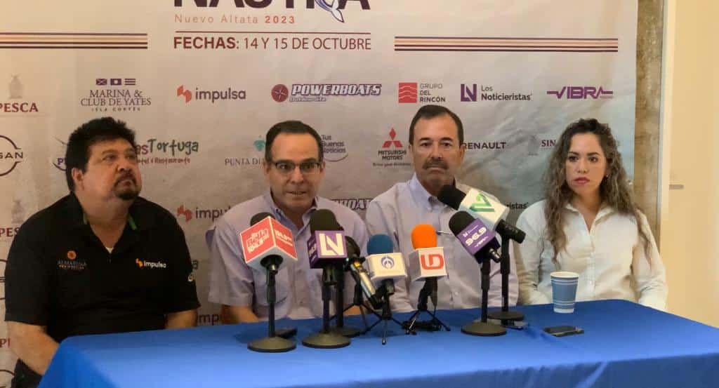 4 personas sentadas, una mesa, micrófonos de medios de comunicación y al fondo una lona con publicidad