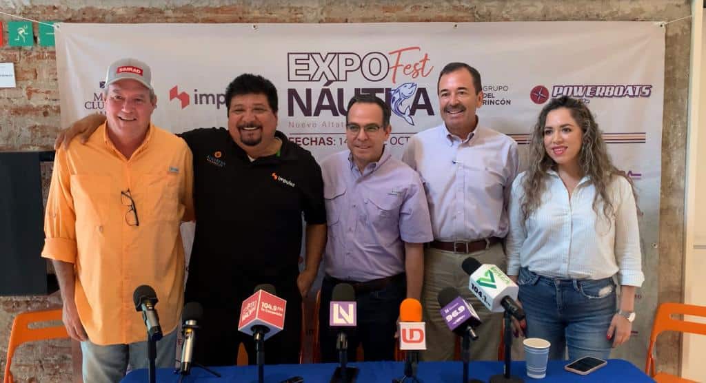 5 personas paradas posando para una foto, una mesa, micrófonos de medios de comunicación y al fondo una lona con publicidad