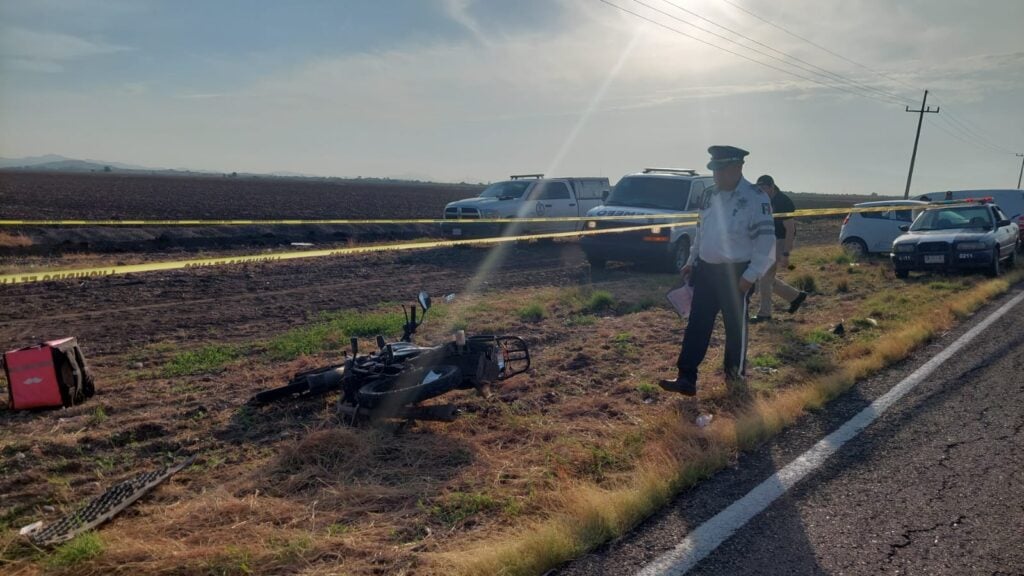 Un persona mirando una motocicleta en un accidente, cinta amarilla delimitando el área y camionetas al fondo