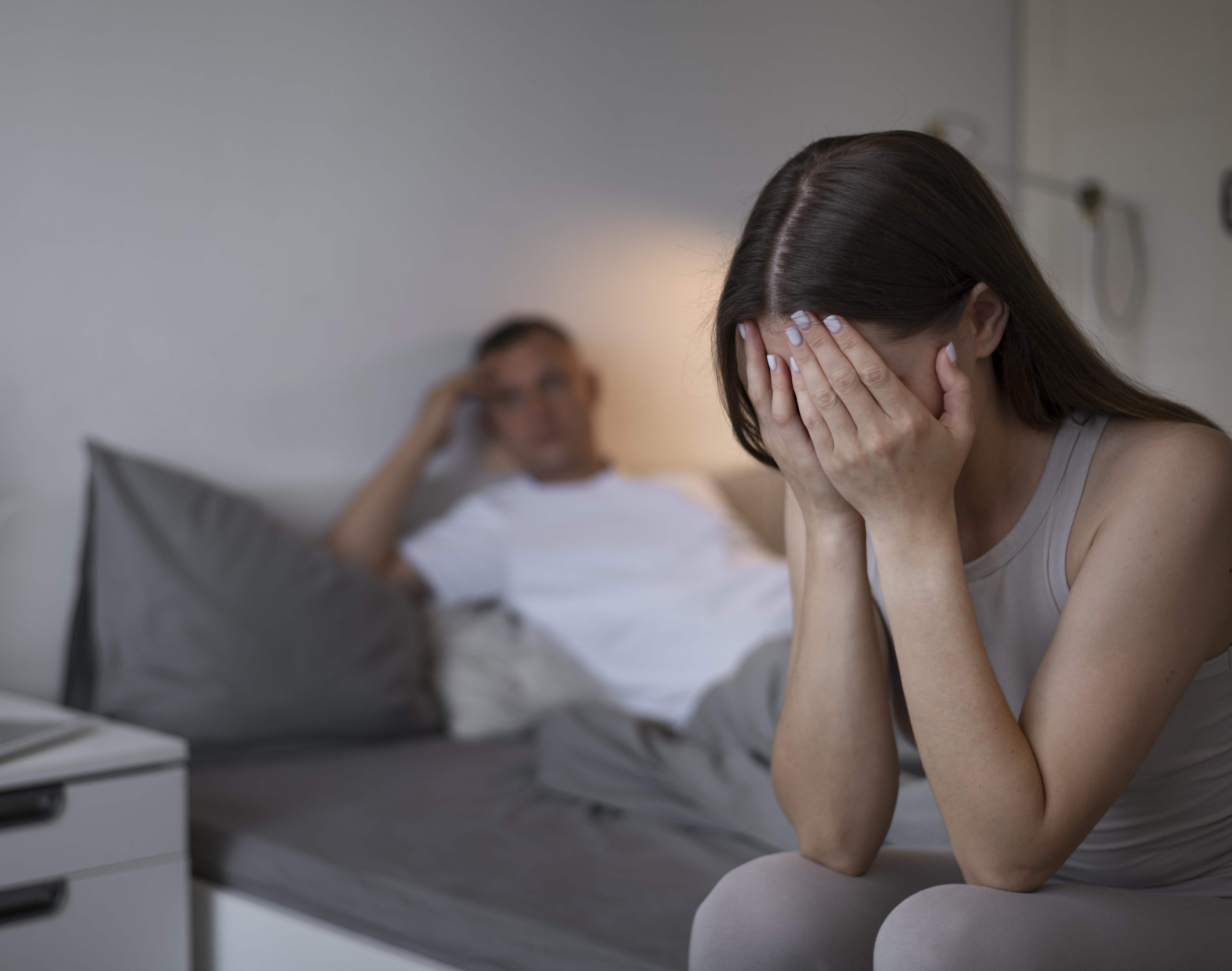 Mujer tapándose la cara con sus manos y hombre acostado en una cama