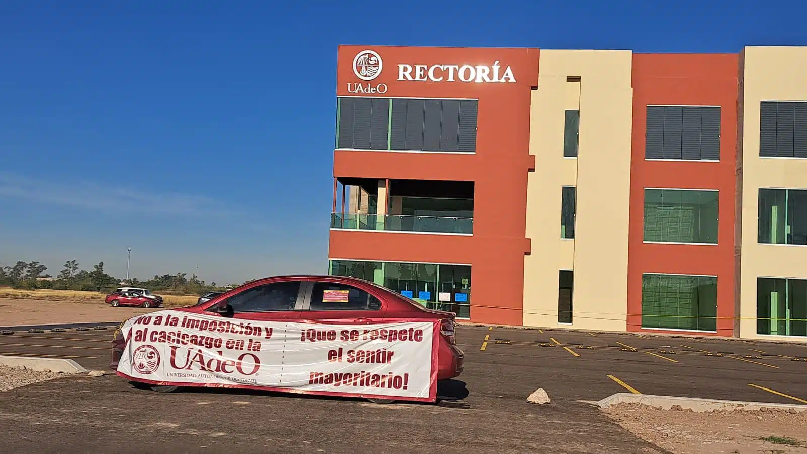 Edificio de Rectoría de la UAdeO y un automóvil con un banner que contiene un mensaje de protesta