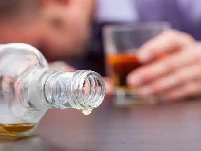 Botella de alcohol derribada sobre una mesa. Al fondo un hombre en estado de ebriedad con un vaso