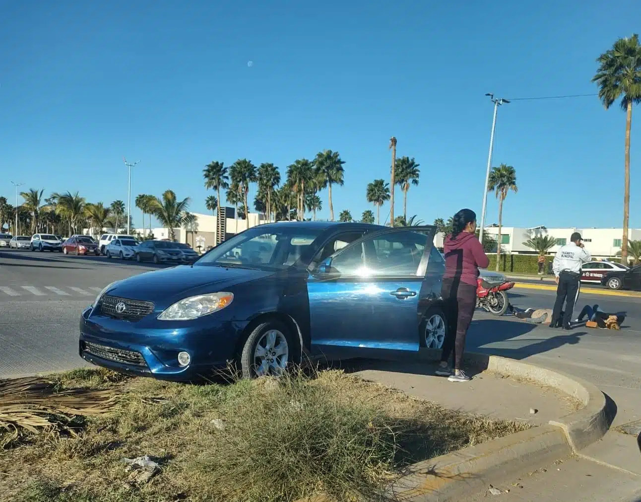 Vehículo color azul chocado sobre una calle de Los Mochis y al fondo una pareja de motociclistas sobre el pavimento