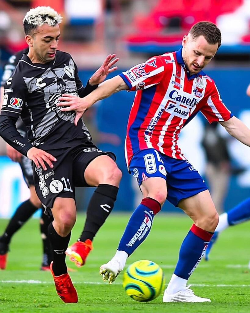 2 jugadores de futbol, uno del equipo de Xolos de Tijuana y el otro del Atlético de San Luis, en una cancha y con un balón