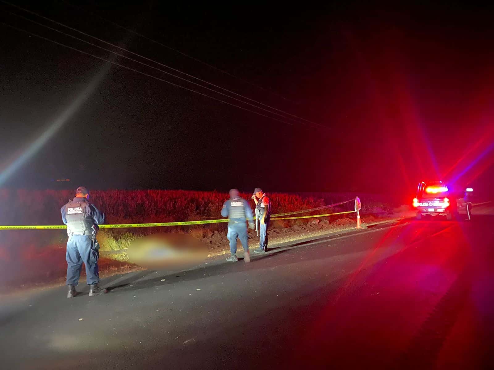 Zona acordonada con cinta amarilla a orilla de una carretera en medio de la noche. Una patrulla y policías también están en la zona