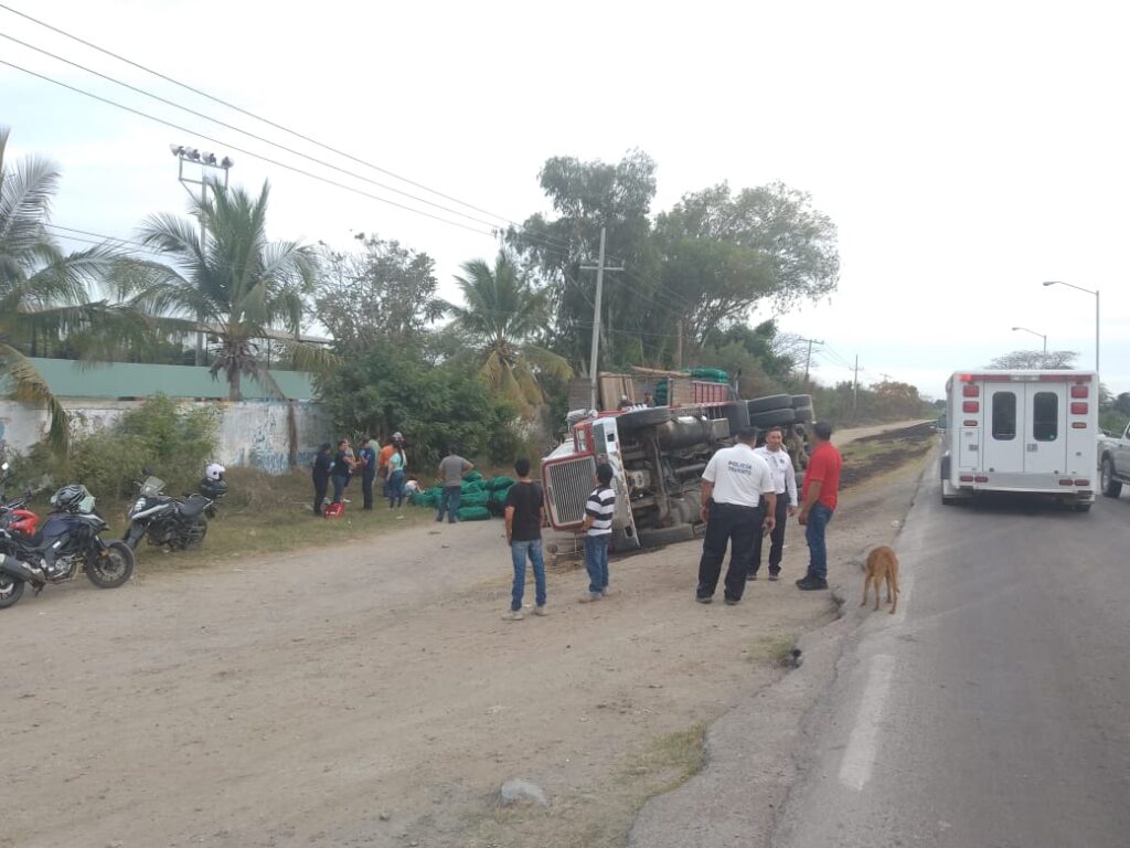 Personas en el lugar donde un tráiler tipo torton cargado de chiles terminó volcado sobre el lado del copiloto en Villa Unión