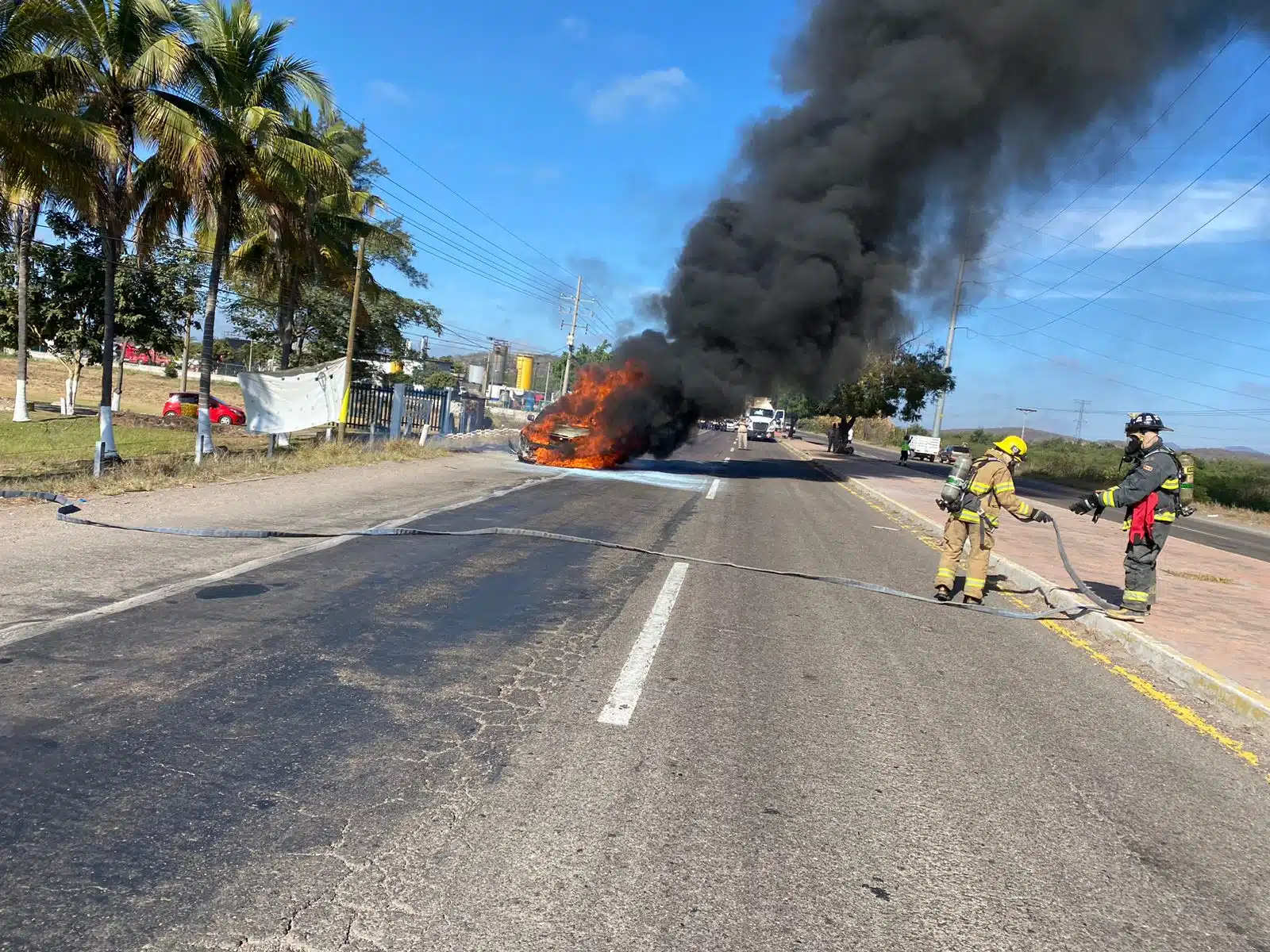 Camioneta incendiándose en una carretera de Mazatlán mientras bomberos se preparan para apagar el fuego