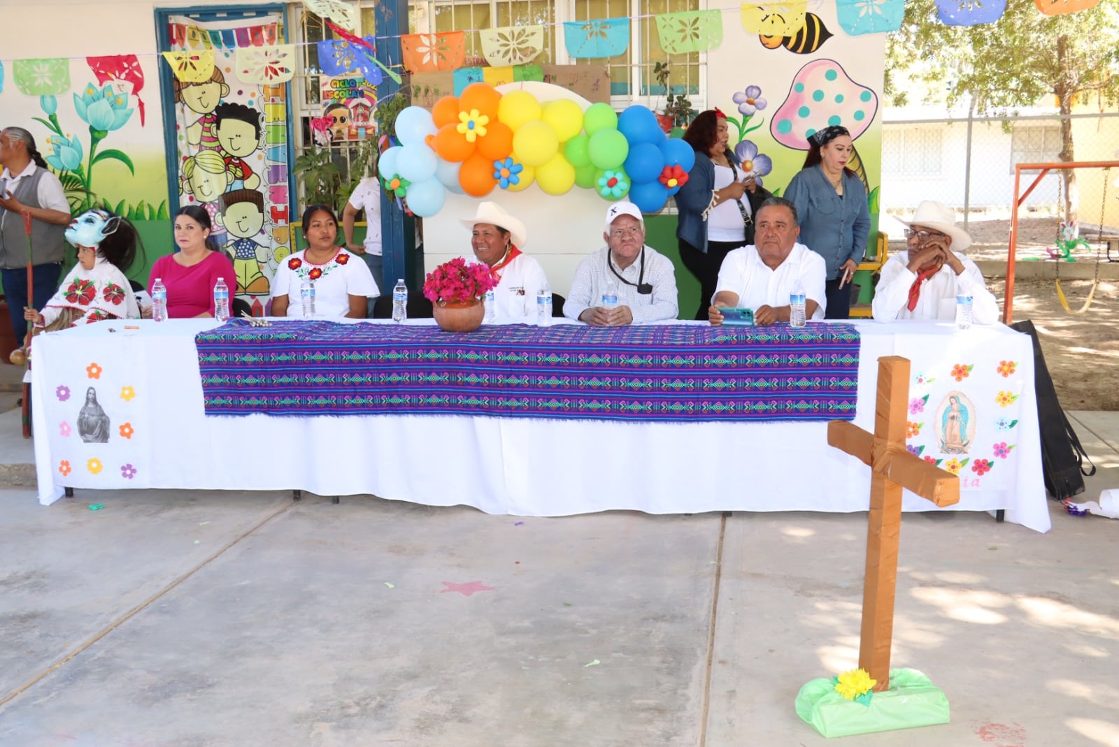 Personas en la mesa del presidium y una cruz de madera en el Jardín de Niños “Genaro Estrada”