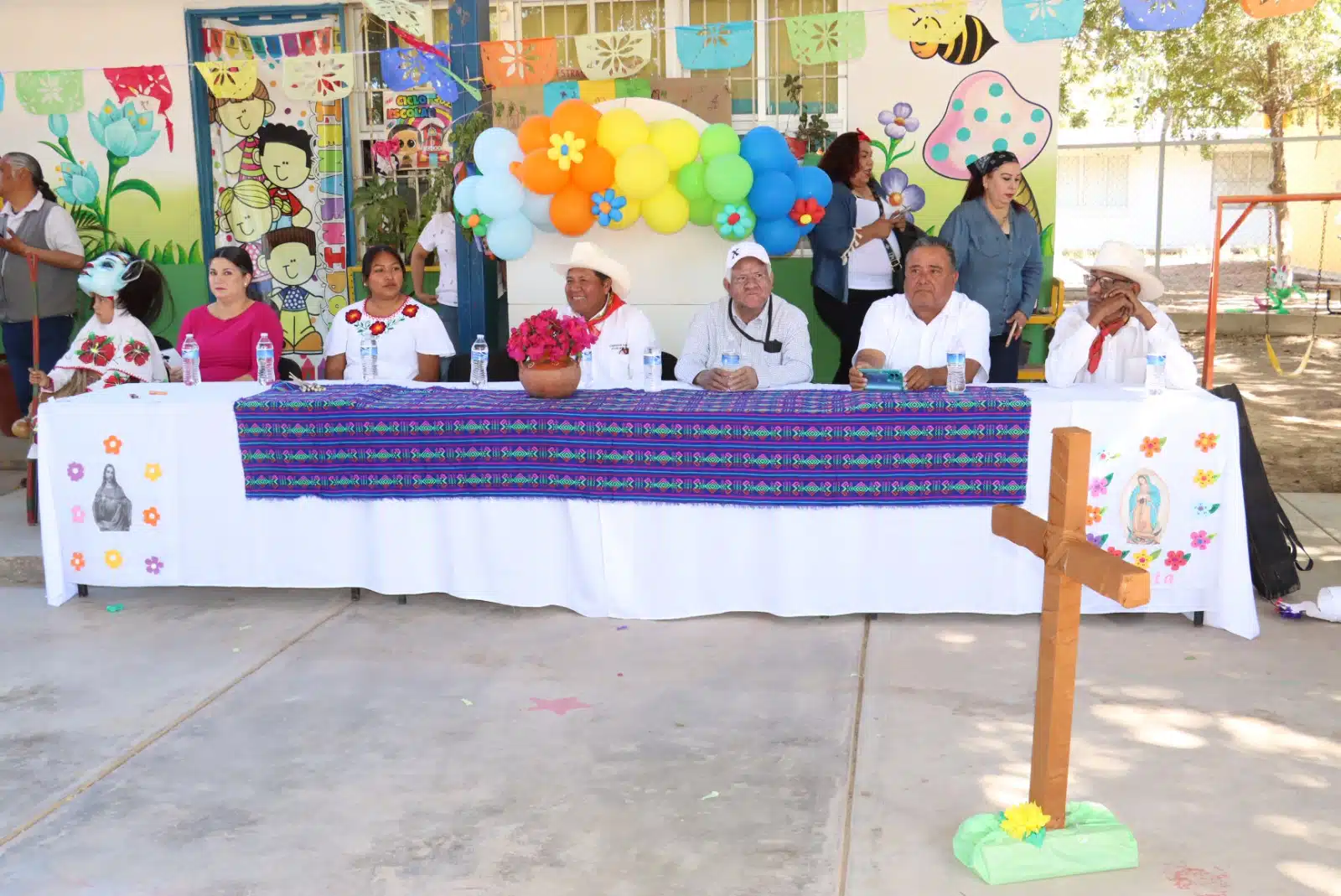 Personas en la mesa del presidium y una cruz de madera en el Jardín de Niños “Genaro Estrada”