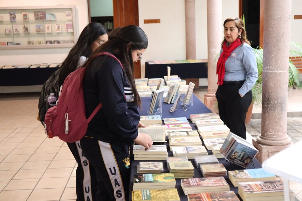 Libros parados y acostados sobre una mesa con un mantel azul, una señora y 2 jovencitas de la UAS en el Colegio Sinaloa en Culiacán