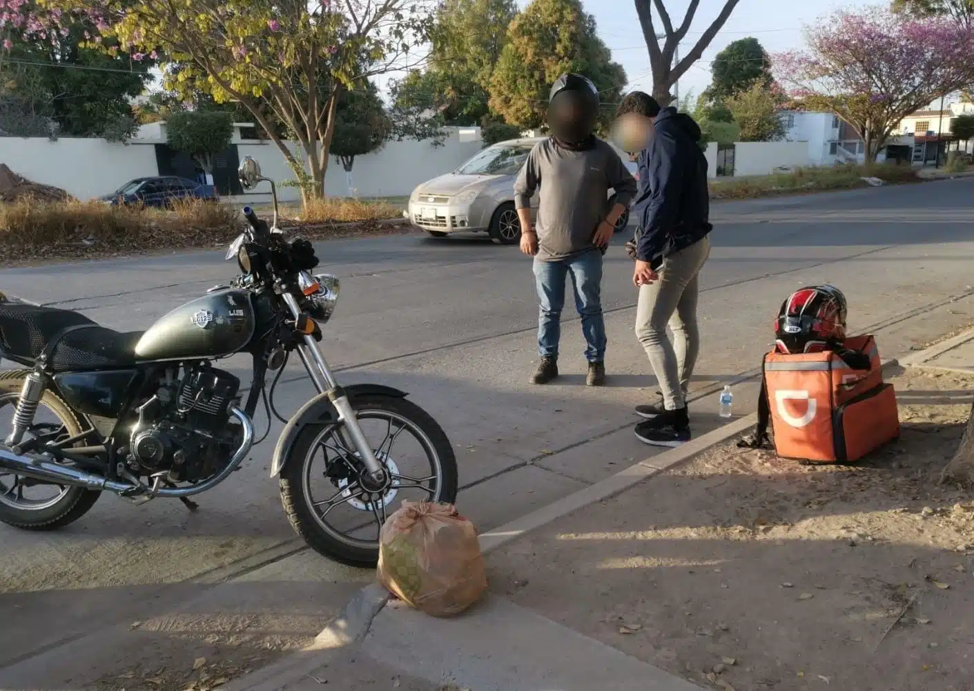 Dos hombres de pie junto a una motocicleta. Al fondo está el carro que participó en el choque