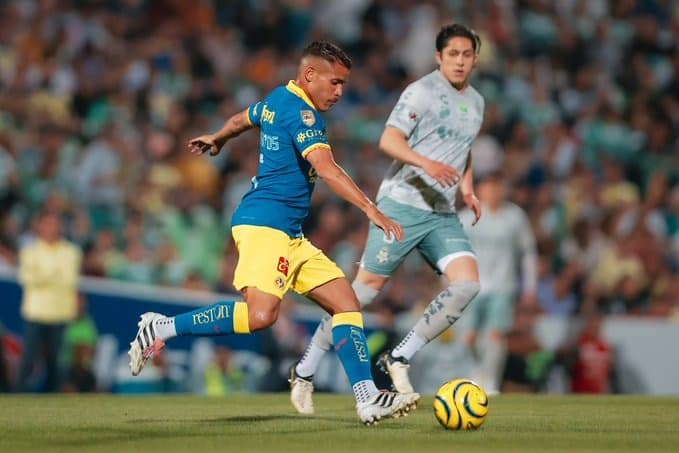 Jonathan Dos Santos jugador del América intentando patear un balón de futbol en una cancha y a un lado un jugador de Santos Laguna