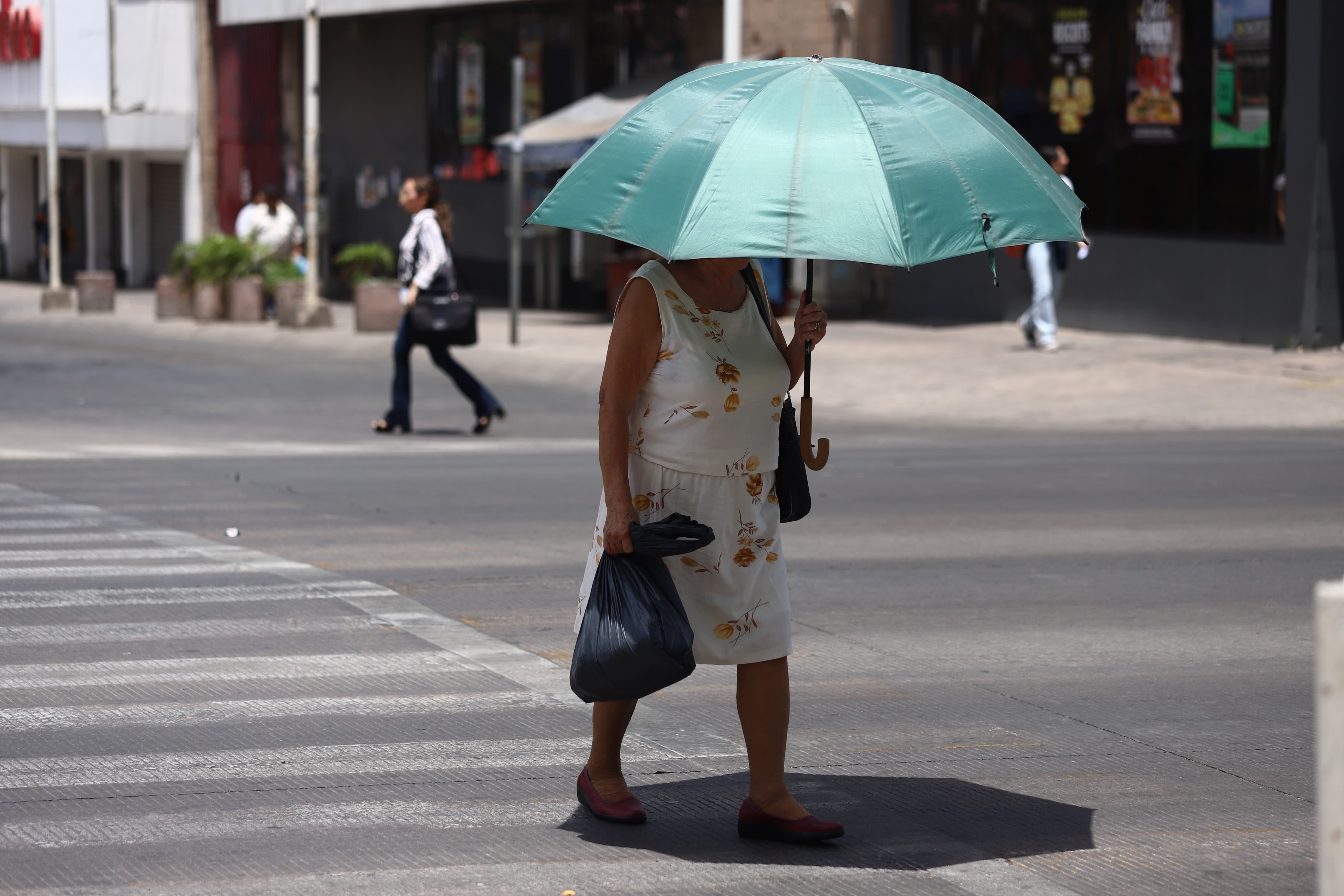 Una señora con un paraguas cubriéndose del sol y caminando por una calle del centro de Culiacán