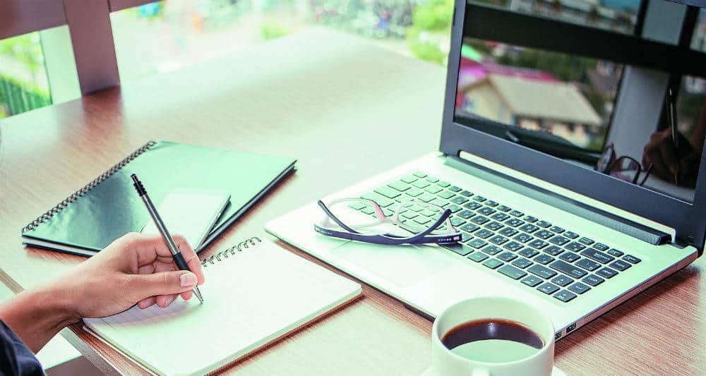 Persona simulando escribir en una libreta con una computadora al frente