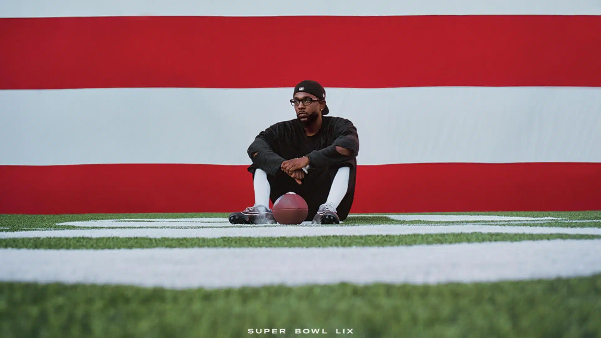 Kendrick Lamar sentado en el pasto y un balón de futbol en sus pies, y detrás suyo, una bandera gigante de Estados Unidos