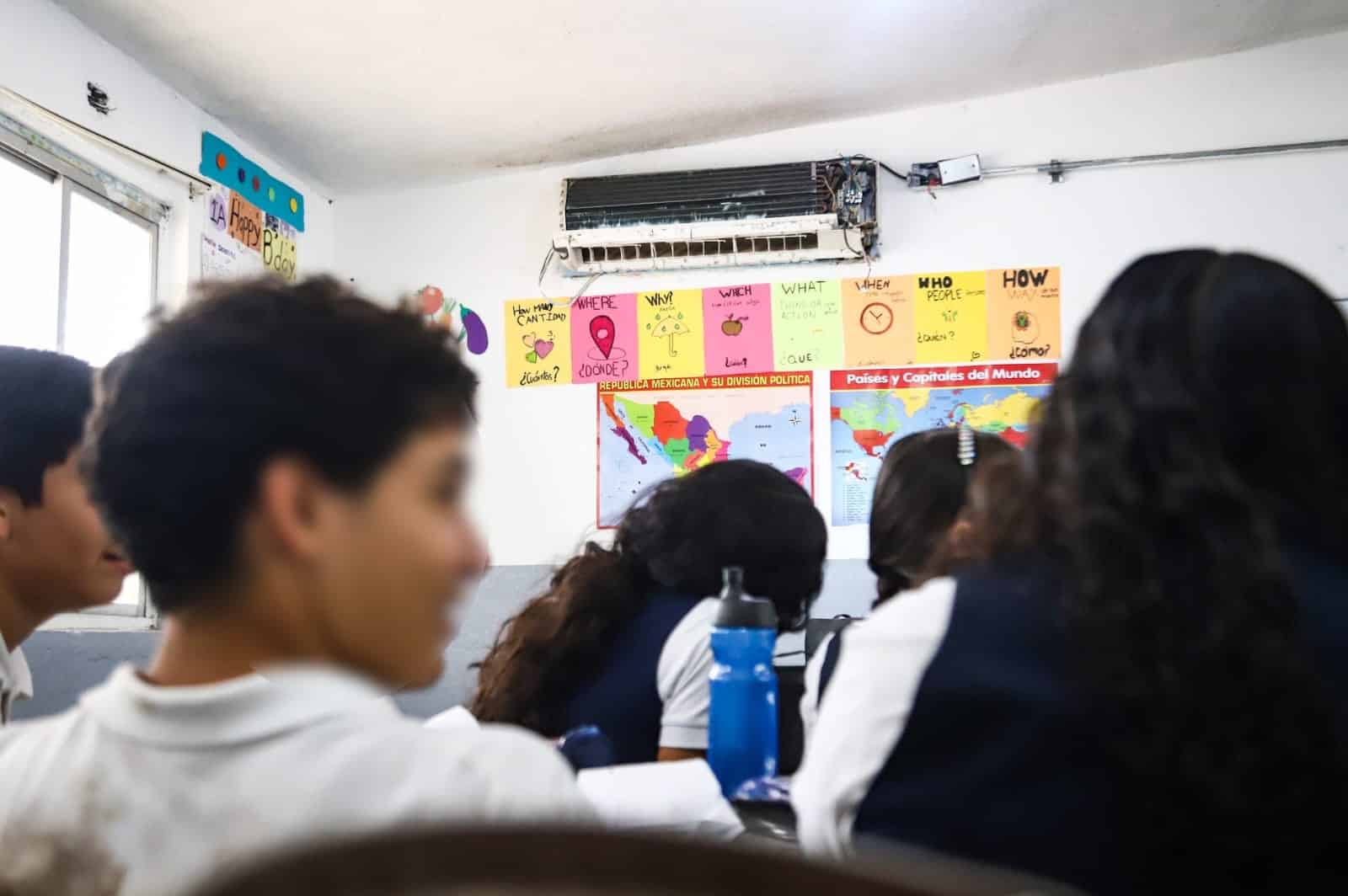 Alumnos en un salón de clases y al fondo un aire acondicionado en malas condiciones