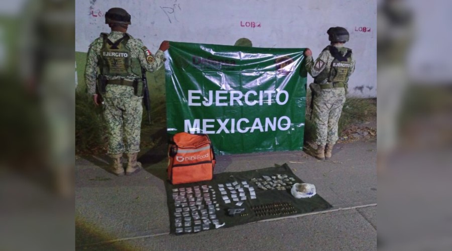 Dos elementos del ejercito mexicano de espaldas sosteniendo una lona con la leyenda del ejercito, y por debajo de ellos en el suelo, las armas y las drogas que aseguraron durante el operativo realizado