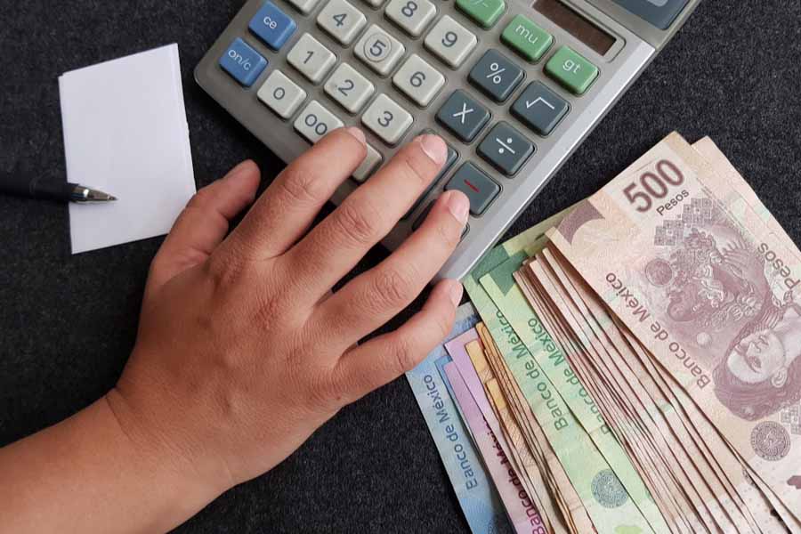 Imagen de billetes mexicanos en sus denominaciones de 500, 200, 100, 50 y 20 pesos, con una calculadora al fondo con papel y pluma y una mano simulando que está contnado el dinero