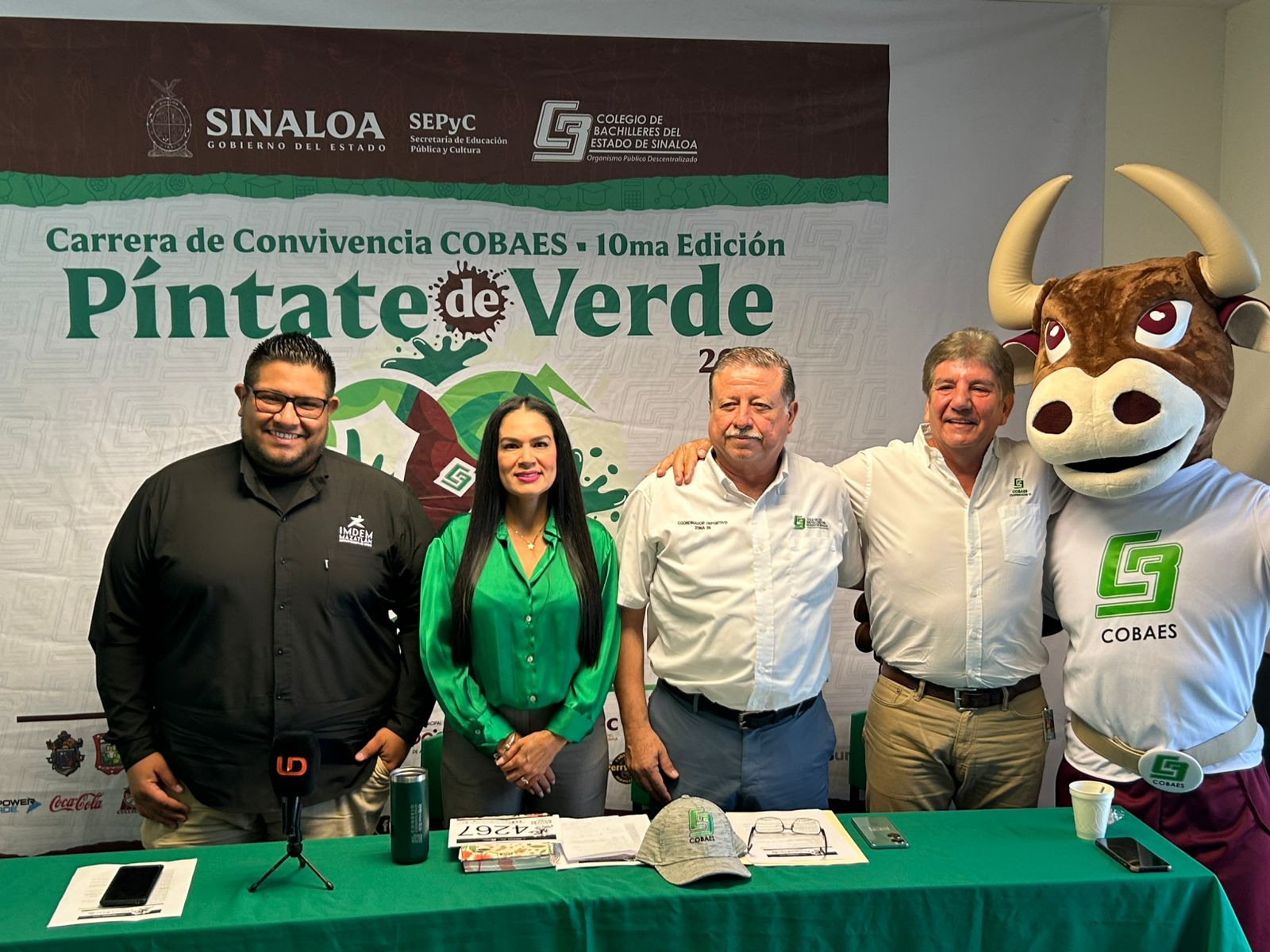 Conferencia de prensa para presentar la carrera de Convivencia Cobaes Píntate de Verde 2025, donde están cuatro autoridades del plantel educativo y a su lado la botarga de la mascota de cobaes, por detrás de ellos el promocional de la carrera.