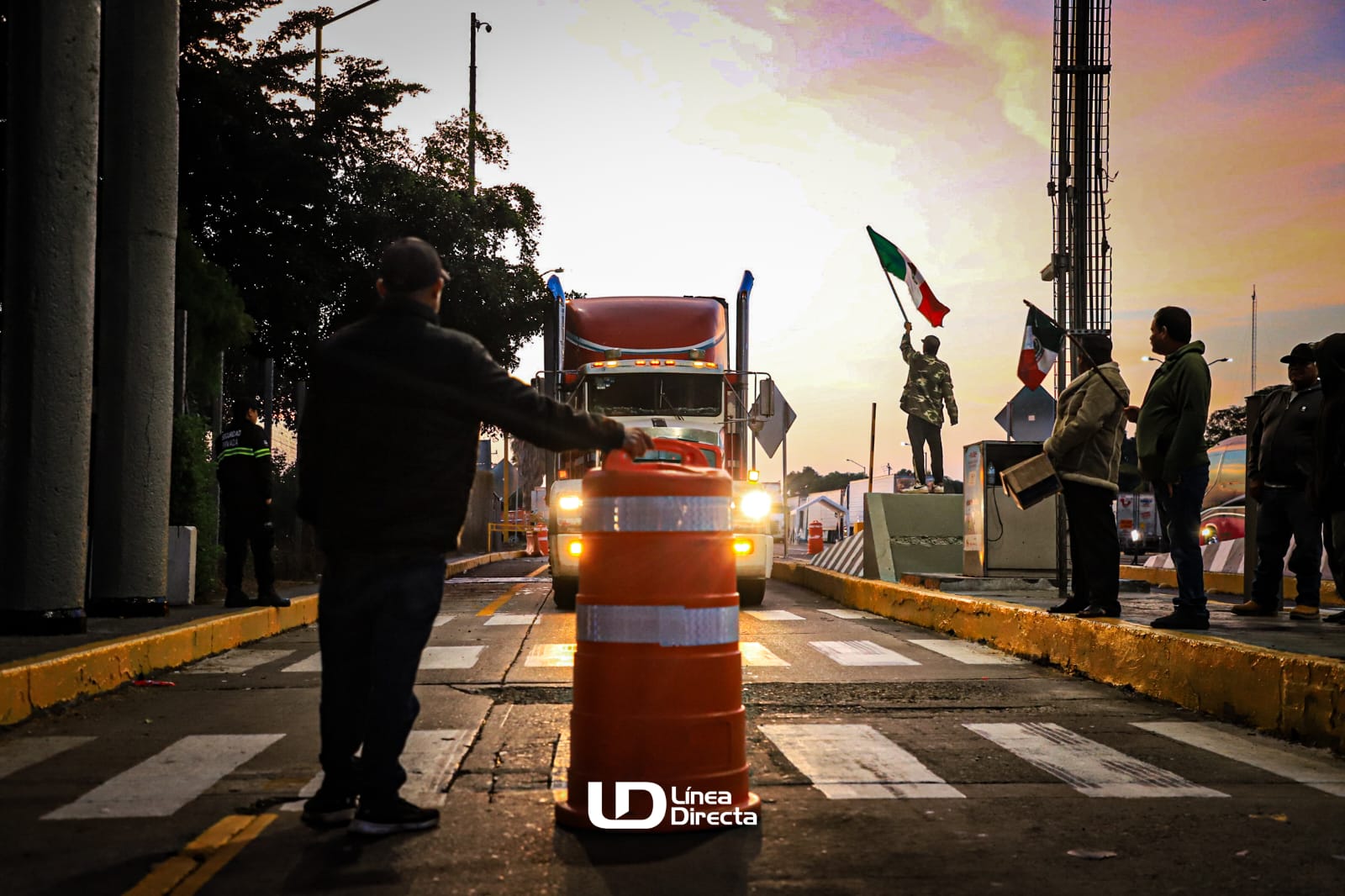 Una persona sostiene un tambo para bloquear el paso junto a una persona con la bandera de México