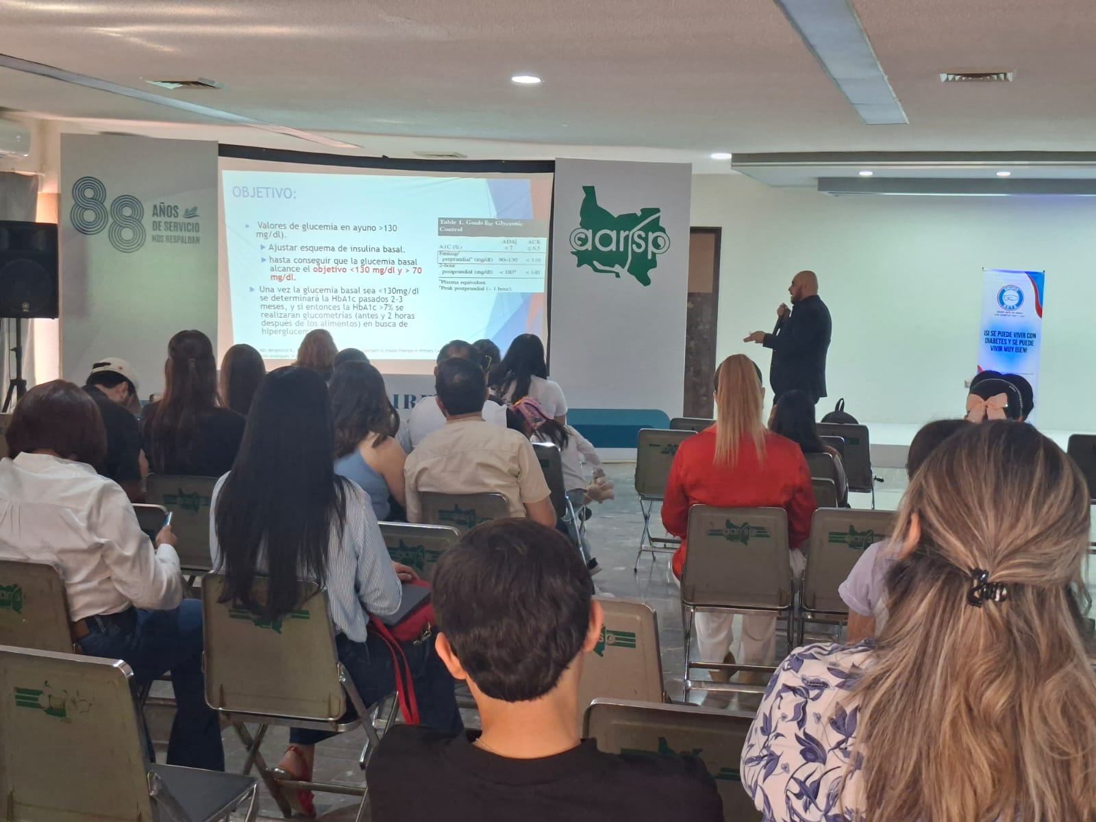 Un grupo de personas durante una charla medica sobre la diabetes, al fondo se observa al ponente y las diapositivas del tema sobre el que esta exponiendo