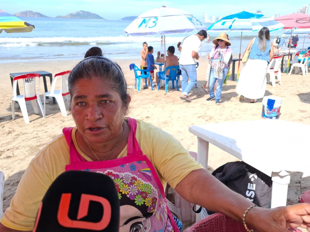 Comerciante de ostiones en Mazatlán debajo de una sombrilla siendo entrevistada, se ve el micrófono de Línea Directa debajo de ella y por detrás turistas y bañistas disfrutando de la playa