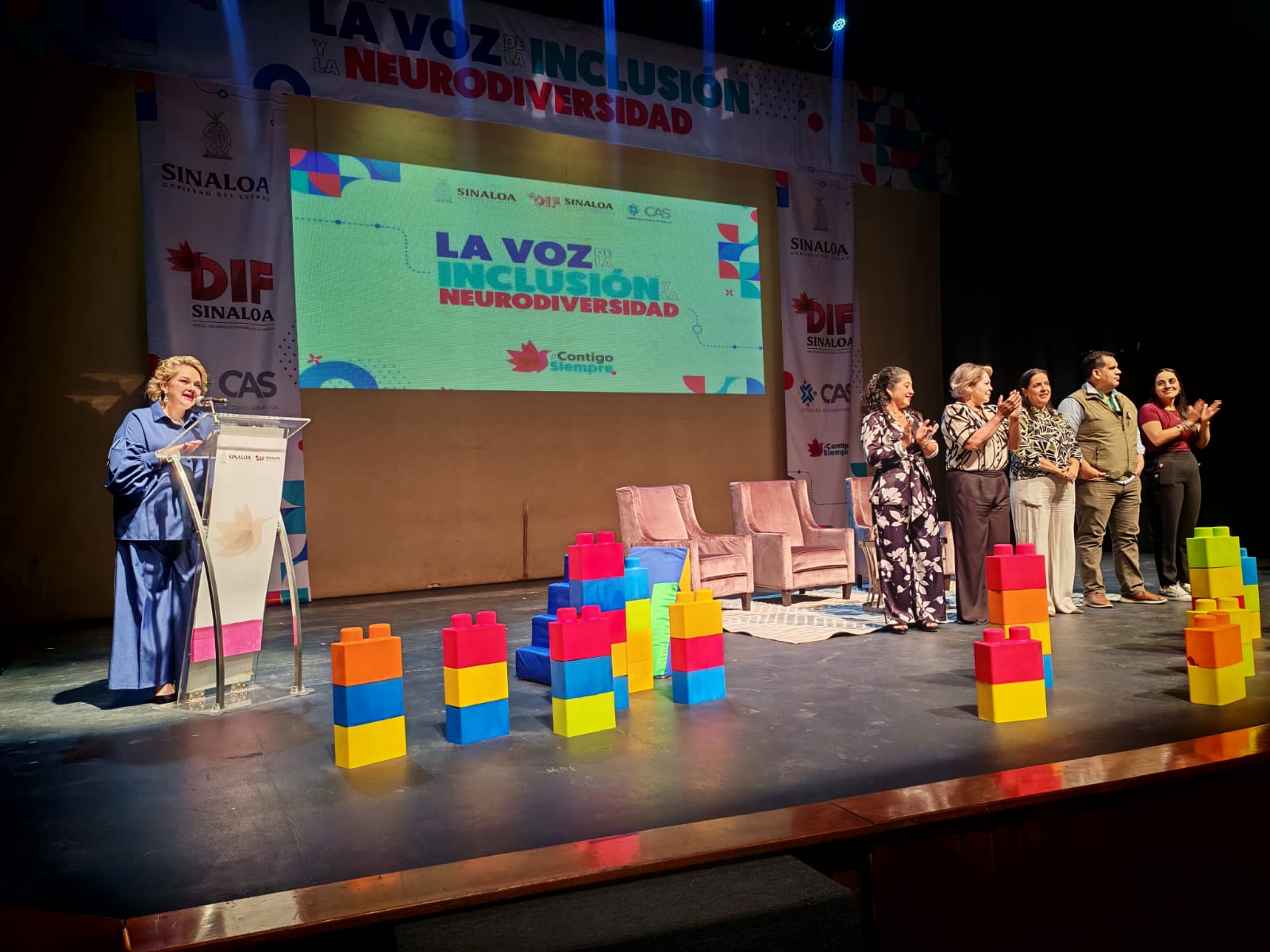 Escenario del Congreso “La Voz de la Inclusión y la Neurodiversidad”, el micrófono la presidenta del DIF Sinaloa, Eneyda Rocha Ruiz y a su lado personas que acompañan en el congreso, se ve decoración alusiva a la temática y proyección de presentaciones.